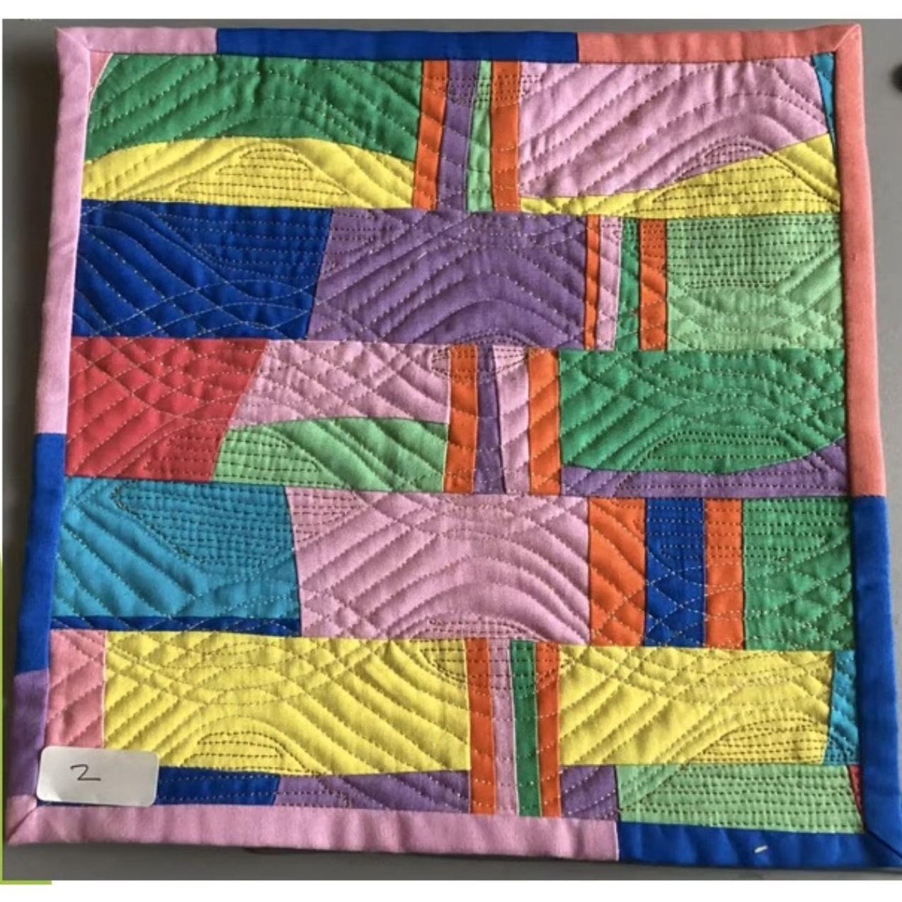 A square quilt with a colorful, abstract design. The quilt features rectangles and triangles in various colors, including pink, green, yellow, blue, and purple. The background is quilted with a linear pattern.