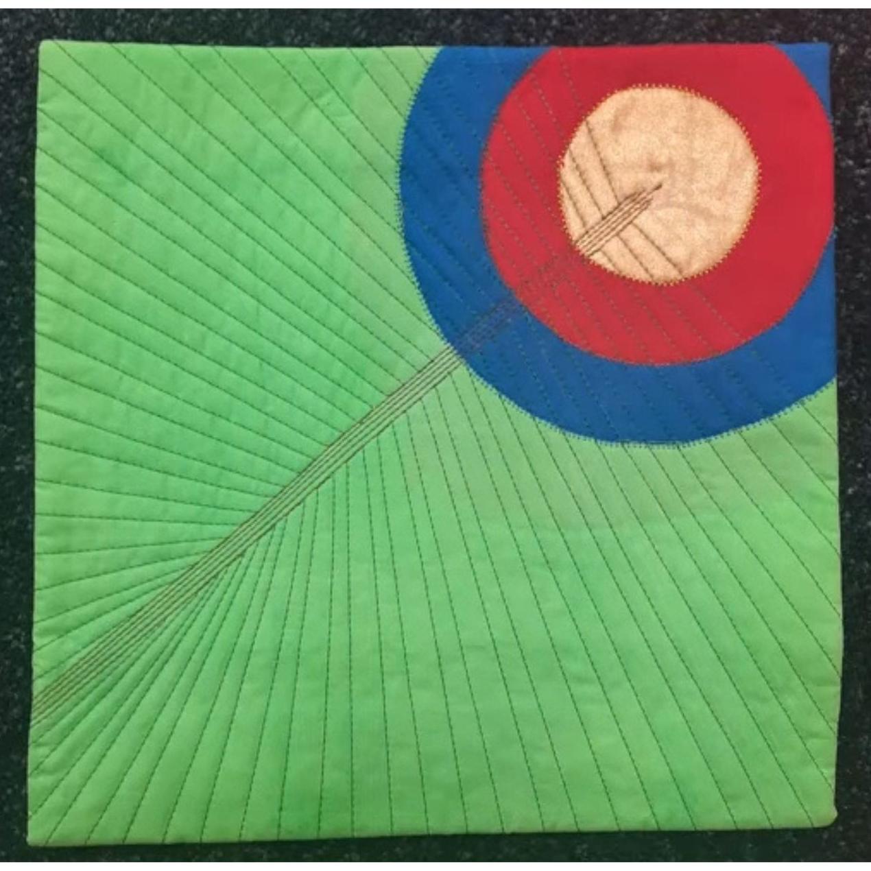 A square quilt with a green background. The quilt features a circular design with concentric circles in red, blue, and white. The background is quilted with radiating lines.