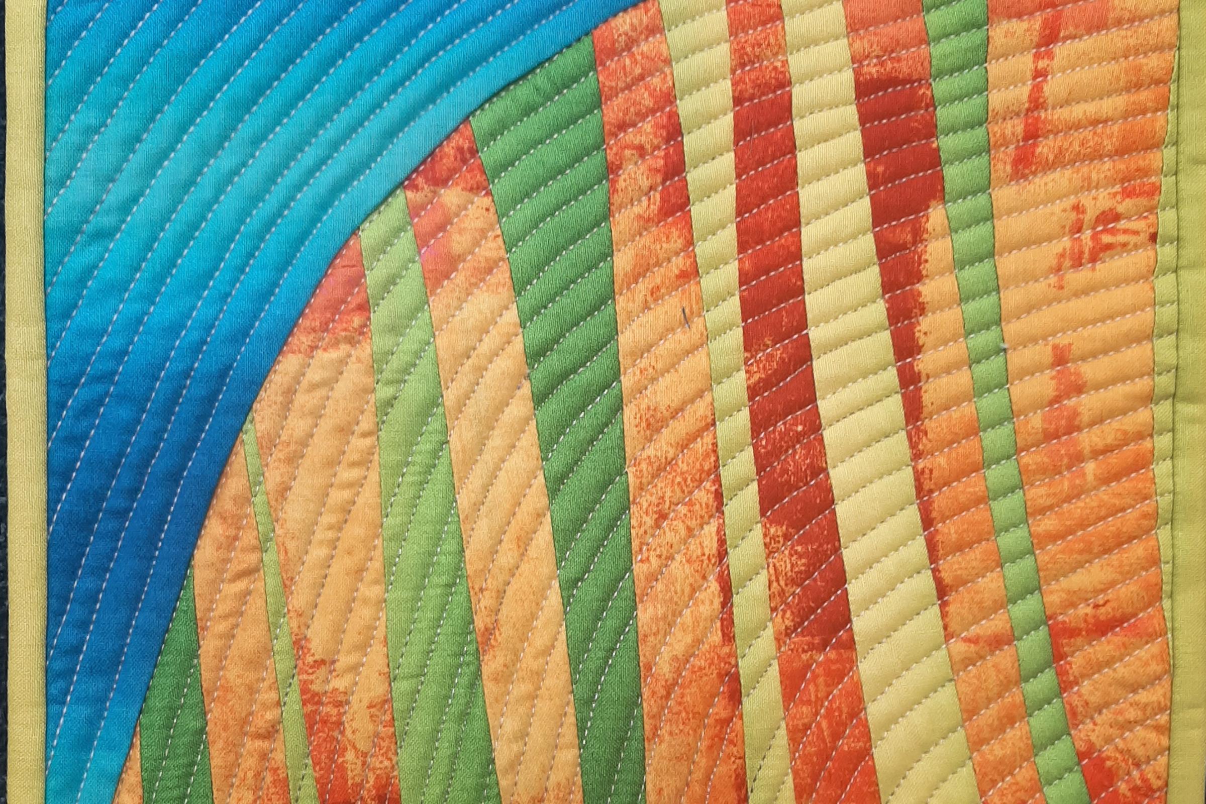 A colourful quilt with a landscape-like design. The quilt features a curved blue section representing the sky, transitioning into vertical strips of green, yellow, and orange, resembling fields or hills. The fabric has a textured, almost painted look, wit