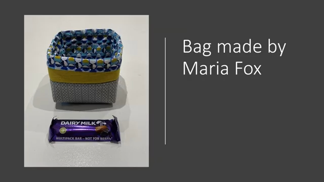 A square, fabric basket. The basket is grey with a yellow band and a patterned blue, yellow and white fabric for the lining.. A Dairy Milk chocolate bar is in front of the basket.