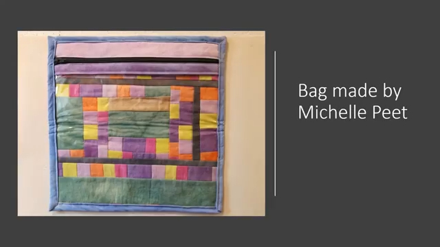 A rectangular, quilted bag with a zipper closure. The bag features a colourful, geometric patchwork design in shades of purple, pink, yellow, green, and grey.