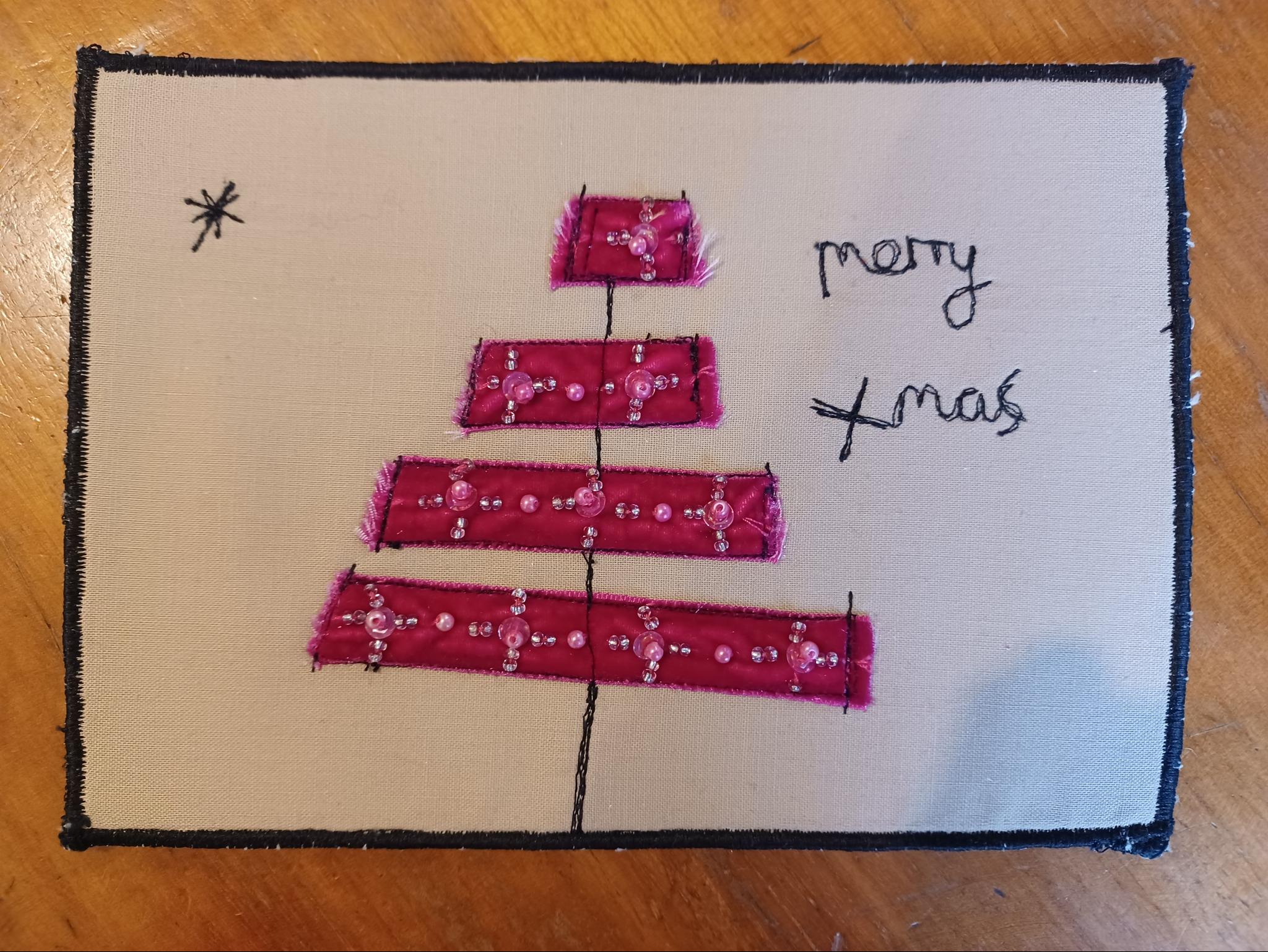 A rectangular quilt with a beige background. The quilt features a pink Christmas tree made up of horizontal strips. The tree is decorated with small, white beads. The words "Merry Xmas" are written in cursive script in the top right corner.