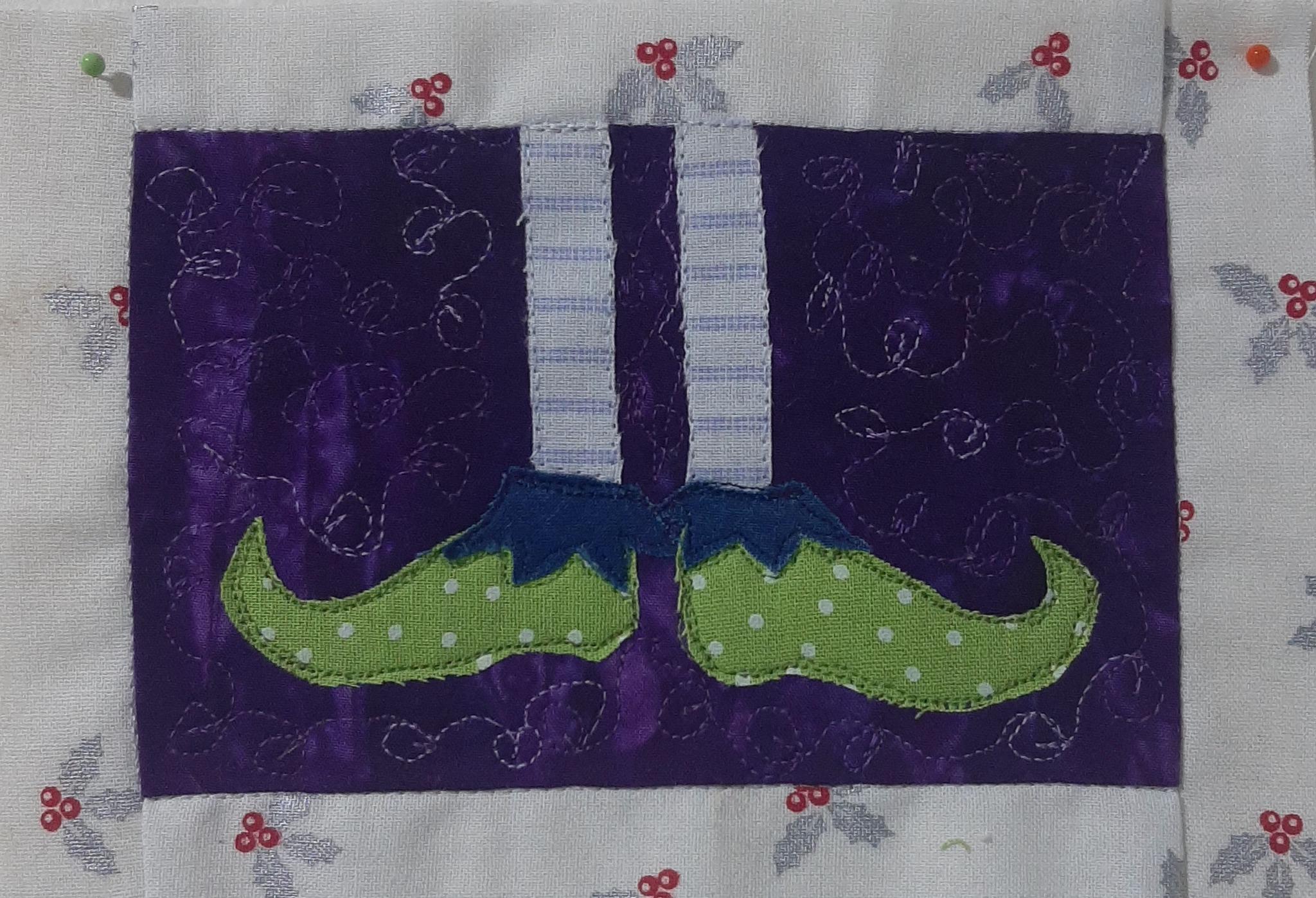 A rectangular quilt with a white background. The quilt features a pair of elf shoes with green polka dot fabric and blue toes. The shoes are on a purple background with a swirling pattern.