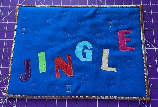 A rectangular quilt with a blue background. The word "JINGLE" is spelled out in large, colourful letters. The background is quilted with a pattern of small, white stars. The quilt has a brown border.
