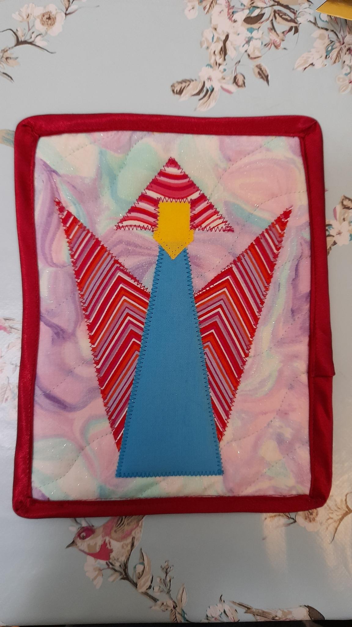 A square quilt with a red border. The quilt features an angel shape made up of colourful triangles, including red, pink, and blue. The angel has a yellow head and a striped body. The background is a light pink with a marbled pattern.