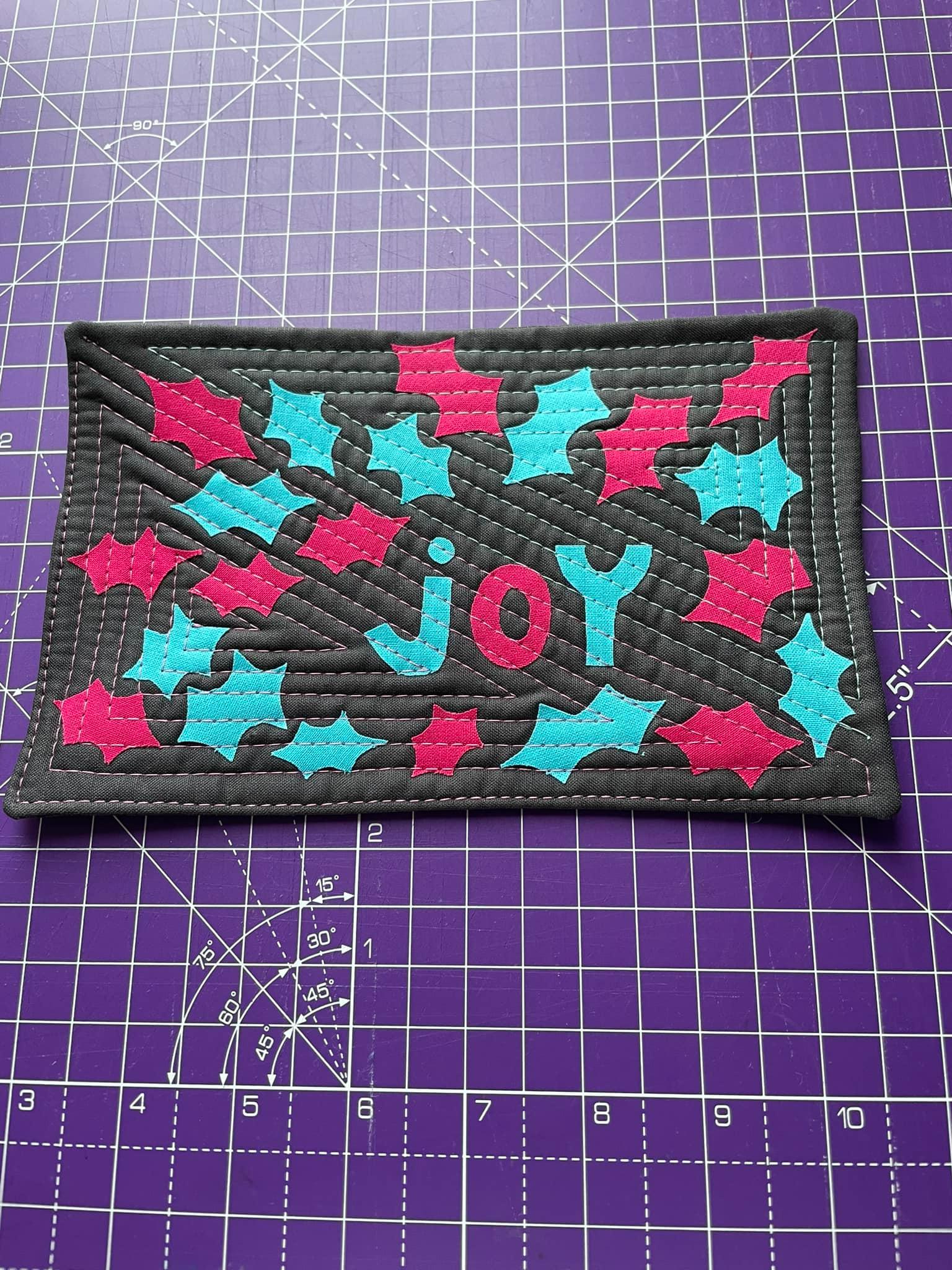 A rectangular quilt with a black background. The word "JOY" is spelled out in large, colourful letters, with pink and blue letters. The background is quilted with a triangular line pattern.
