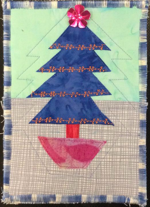 A square quilt with a green background. The quilt features a blue Christmas tree with a pink base and a pink flower on top. The background is quilted with a grid pattern. The quilt has a blue border.