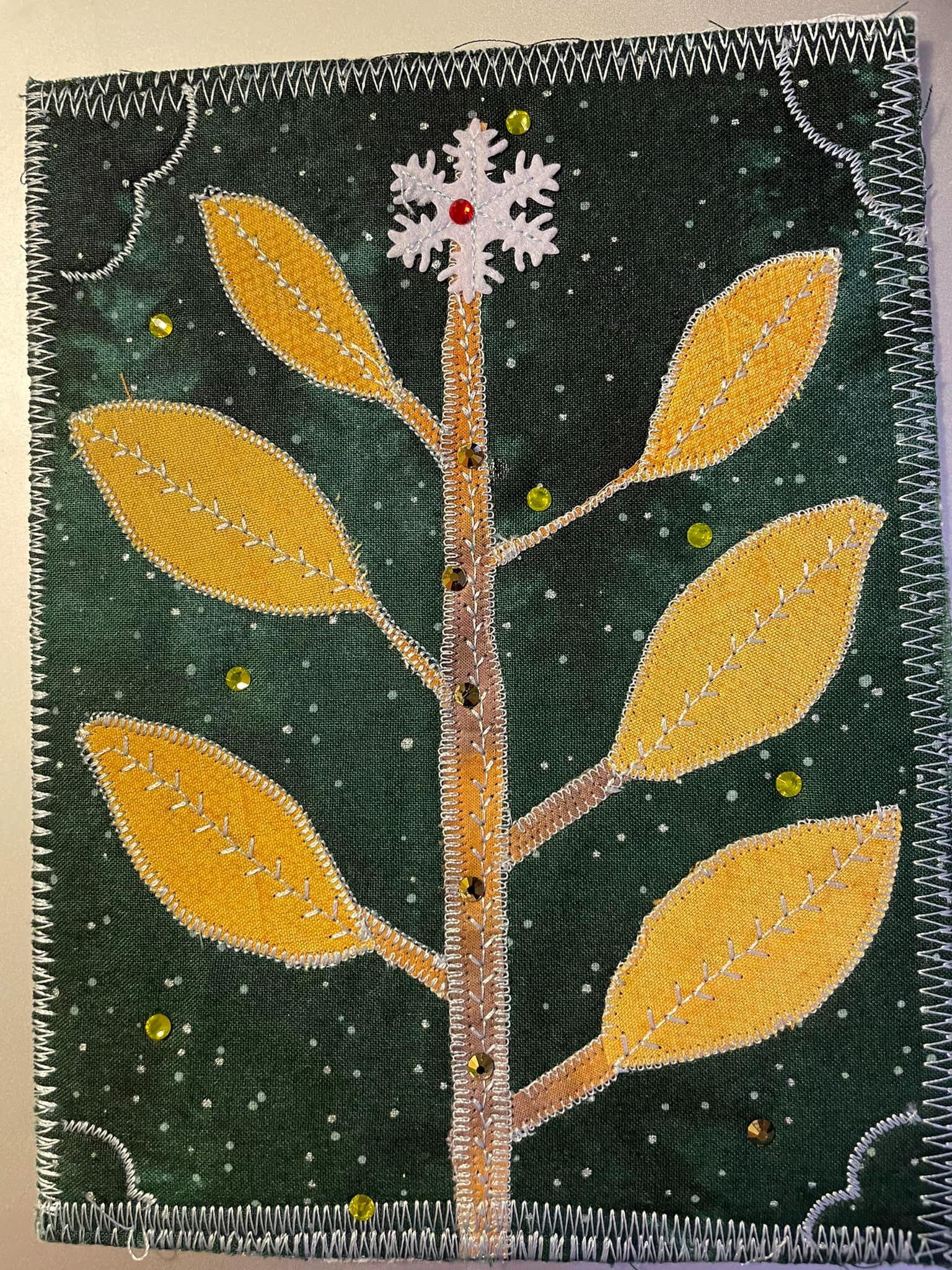 A square quilt with a dark green background. The quilt features a stylized tree with yellow leaves and a brown trunk. There is a snowflake with a red centre at the top of the tree. The background is filled with small, white dots. The quilt has a white border.