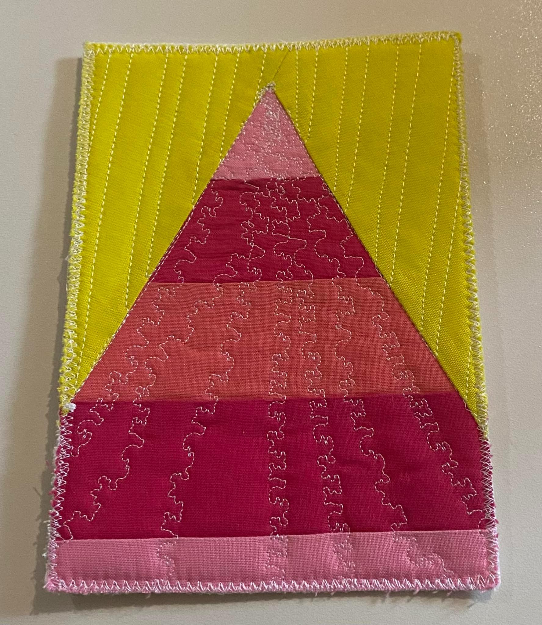 A postcard quilt with a yellow background. The quilt features a large, pink triangle made up of smaller, pink triangles. The background is quilted with vertical lines.