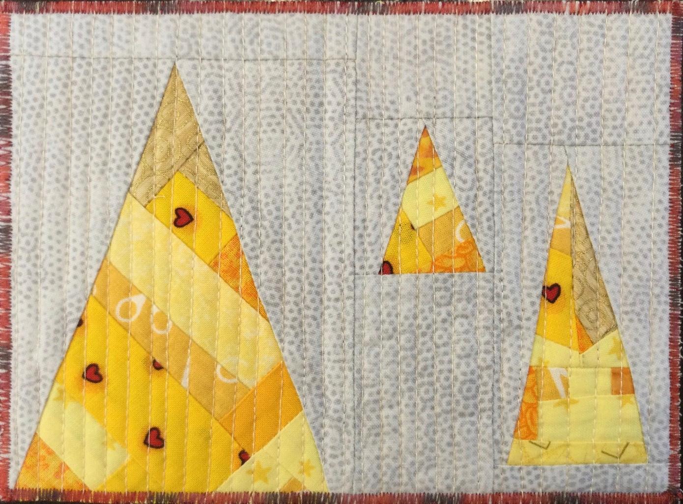 A rectangular quilt with a grey background. The quilt features three yellow triangles of different sizes. The triangles are made up of smaller, colourful triangles and have a textured, quilted appearance. The background is quilted with a subtle pattern of small dots.
