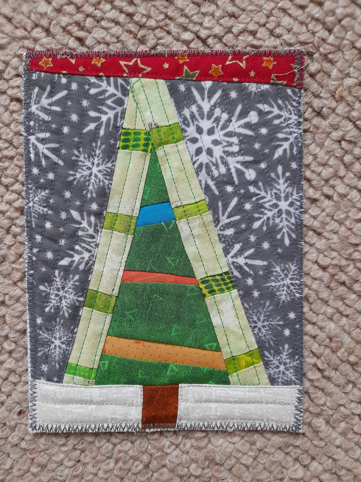 A square quilt with a grey background. The quilt features a colourful Christmas tree made up of strips of fabric in green, orange, red, and blue. The background is filled with white snowflakes.