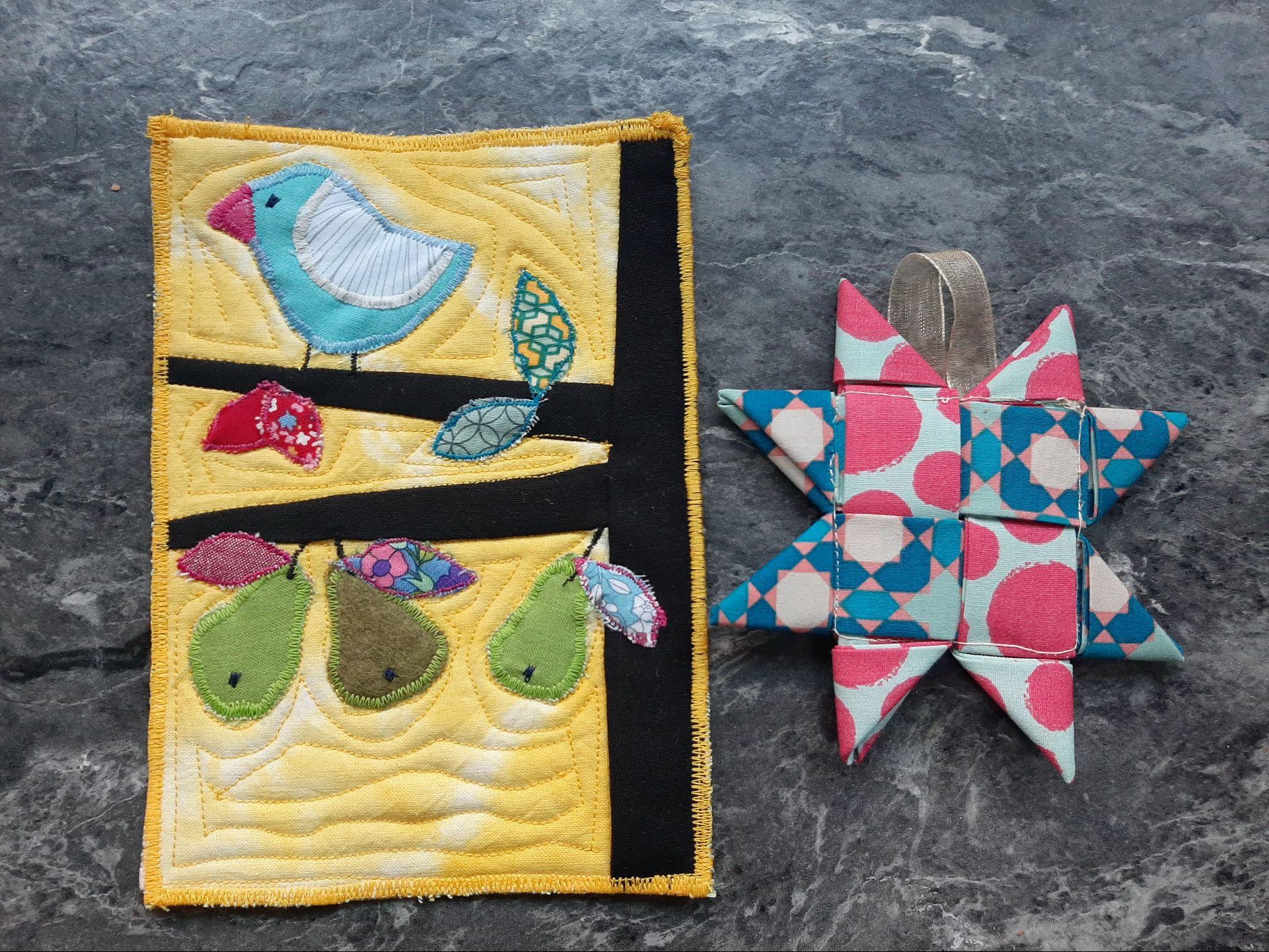 Two small, quilted pieces. The left piece is a rectangular wall hanging with a yellow background. It features a blue bird perched on a branch with colourful pears and leaves. The right piece is a hanging, star-shaped ornament with a patchwork design in blue, pink, and white.