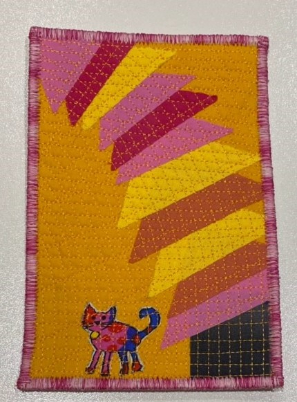 A rectangular quilt with a yellow background. The quilt features a colourful, geometric design with overlapping triangles in shades of pink and orange. In the bottom left corner, there is a small, colourful cat. The quilt has a pink border.