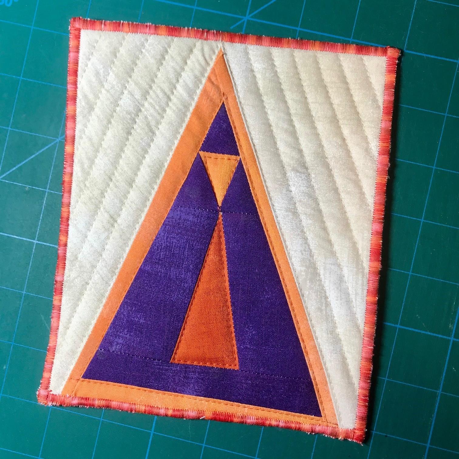 A square quilt with a white background. The quilt features a large, purple triangle with an orange triangle inside. The background is quilted with a linear pattern. The quilt has an orange border.