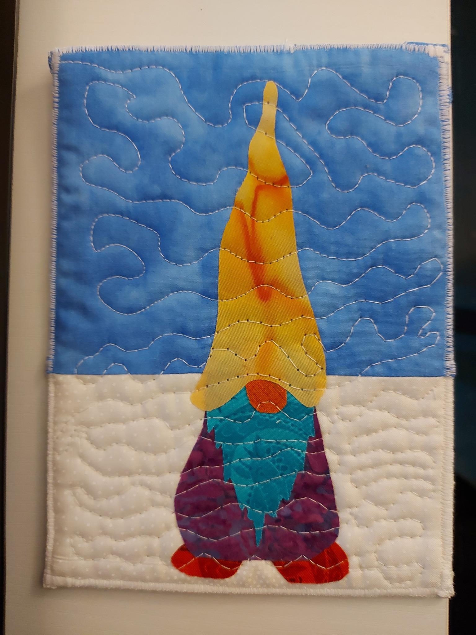 A square quilt with a blue background. The quilt features a gnome with a yellow hat, a purple beard, and red boots. The background is quilted with a wavy pattern representing snow.