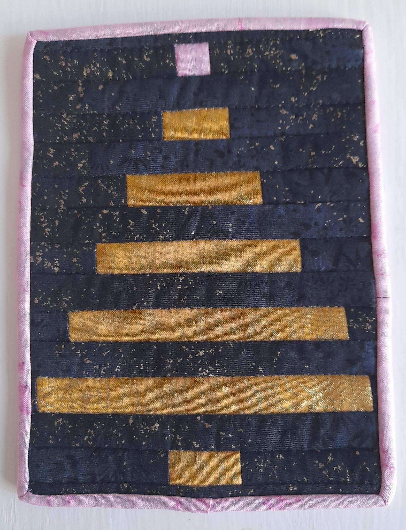 A square quilt with a pink background. The quilt features a Christmas tree made up of horizontal, gold rectangles on a dark, speckled background. The quilt has a pink border.