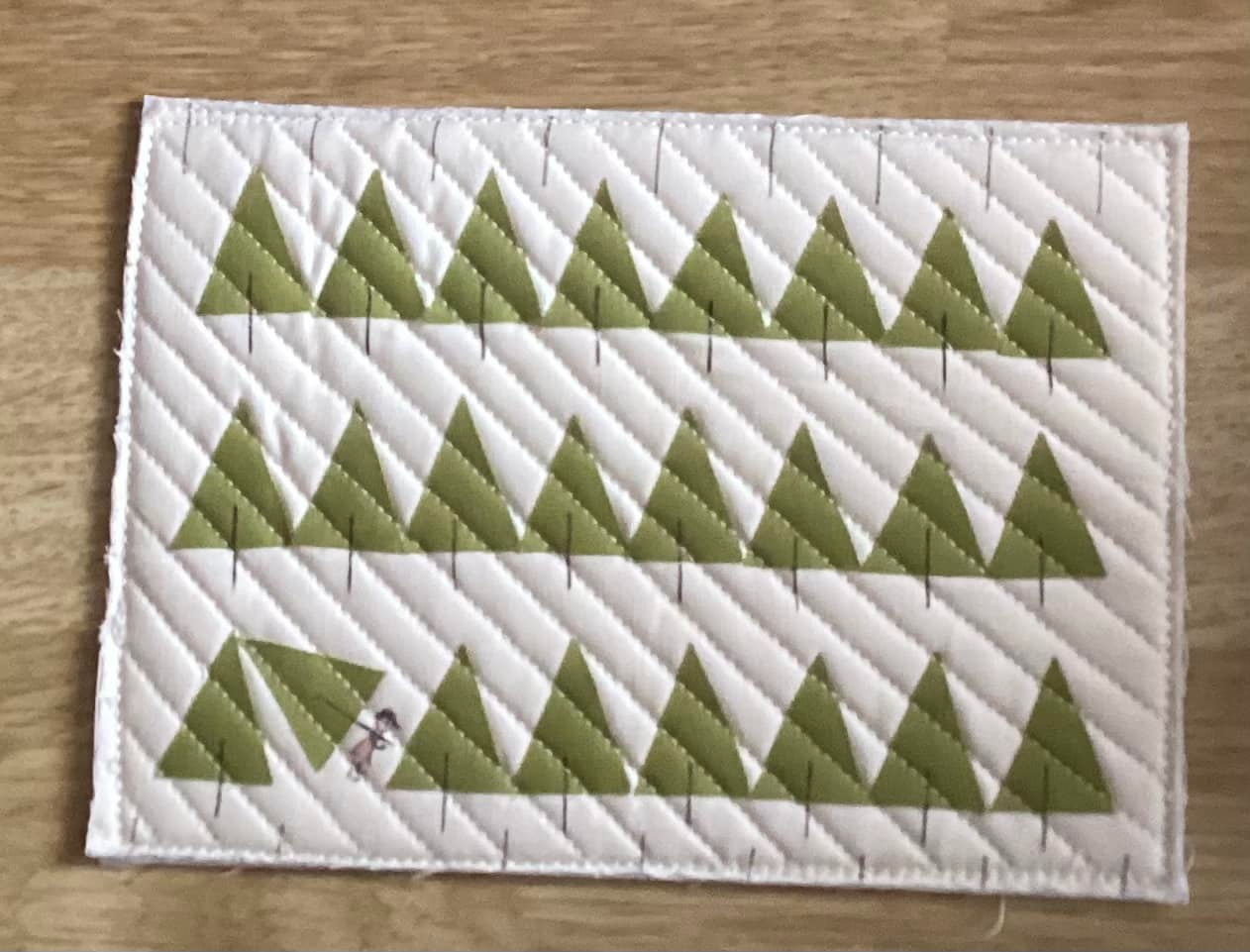 A rectangular quilt with a white background. The quilt features multiple rows of green triangle shapes, representing Christmas trees. One of the trees has a small figure, carrying a chopped down tree. The background is quilted with a diagonal line pattern.