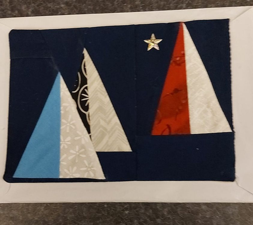 A square quilt with a dark blue background. The quilt features three Christmas trees of different sizes and colours: one blue, one white with a black pattern, and one red and white. A small, gold star is placed at the top of the quilt.
