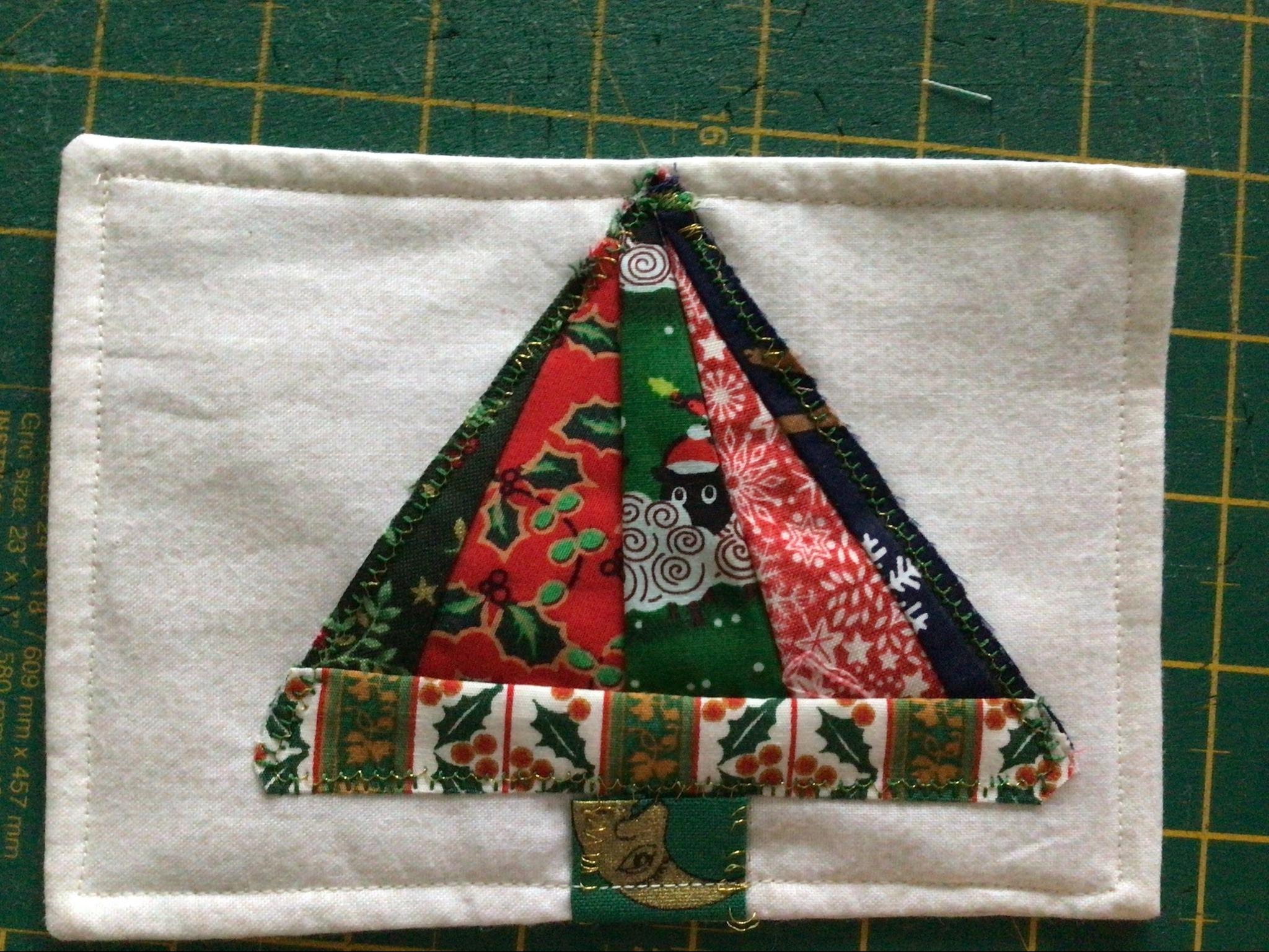 A square quilt with a white background. The quilt features a colourful Christmas tree made up of triangles in various fabrics, including green, red, and blue. The tree is decorated with a small, gold star.