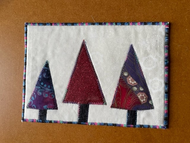 A rectangular quilt with a white background. The quilt features three Christmas trees of different sizes, made of purple and red fabric. The trees have brown trunks. The quilt has a colourful border.
