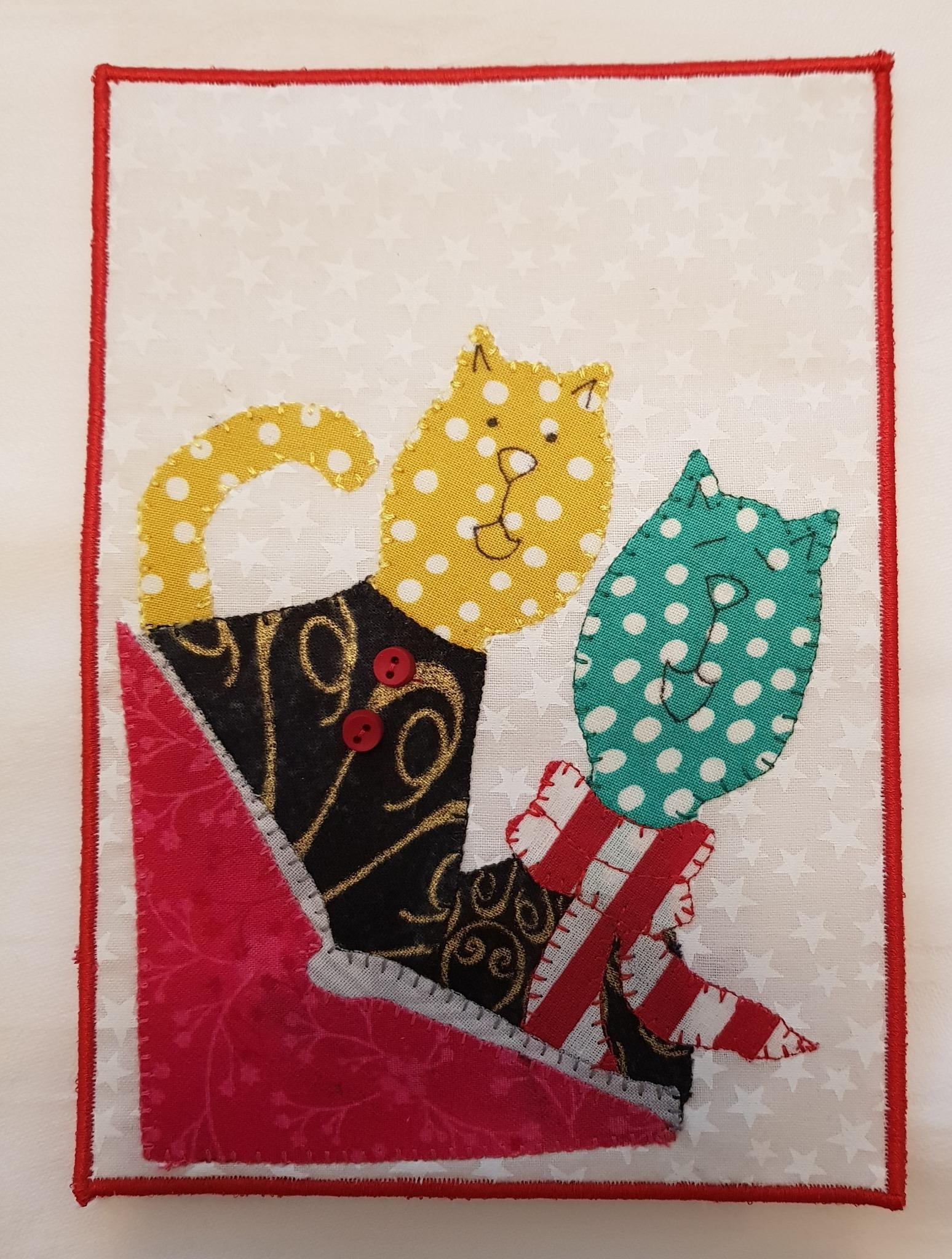 A rectangular quilt with a white background. The quilt features two cats, one yellow and one green, peeking out of a red box. The cats have polka dot patterns and button eyes. The box is decorated with a red and white ribbon. The quilt has a red border.