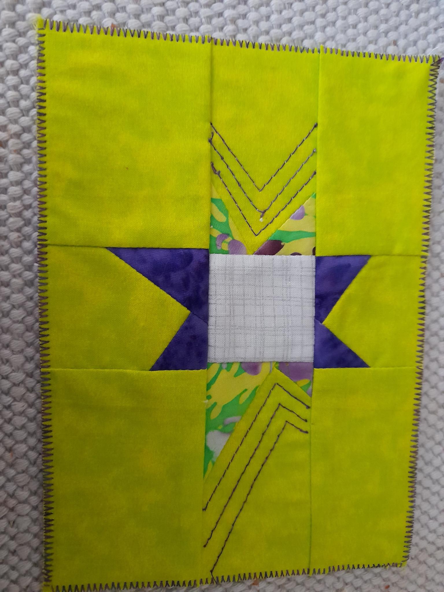 A square quilt with a bright yellow background. The quilt features a star-shaped design with purple triangles and a white centre.