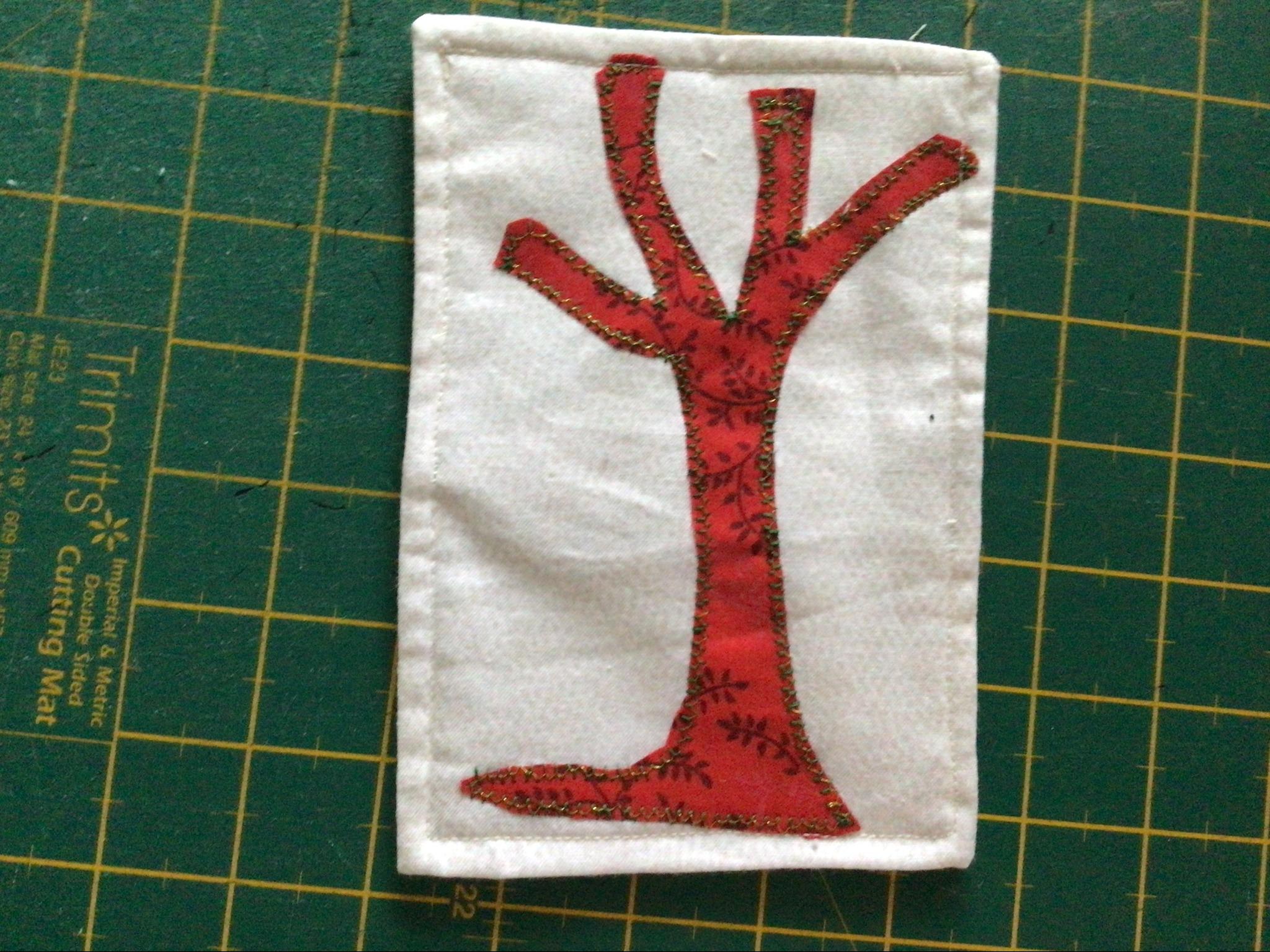A rectangular quilt with a white background. The quilt features a red tree with a gold glitter pattern on the trunk and branches. The quilt is resting on a green cutting mat with markings.