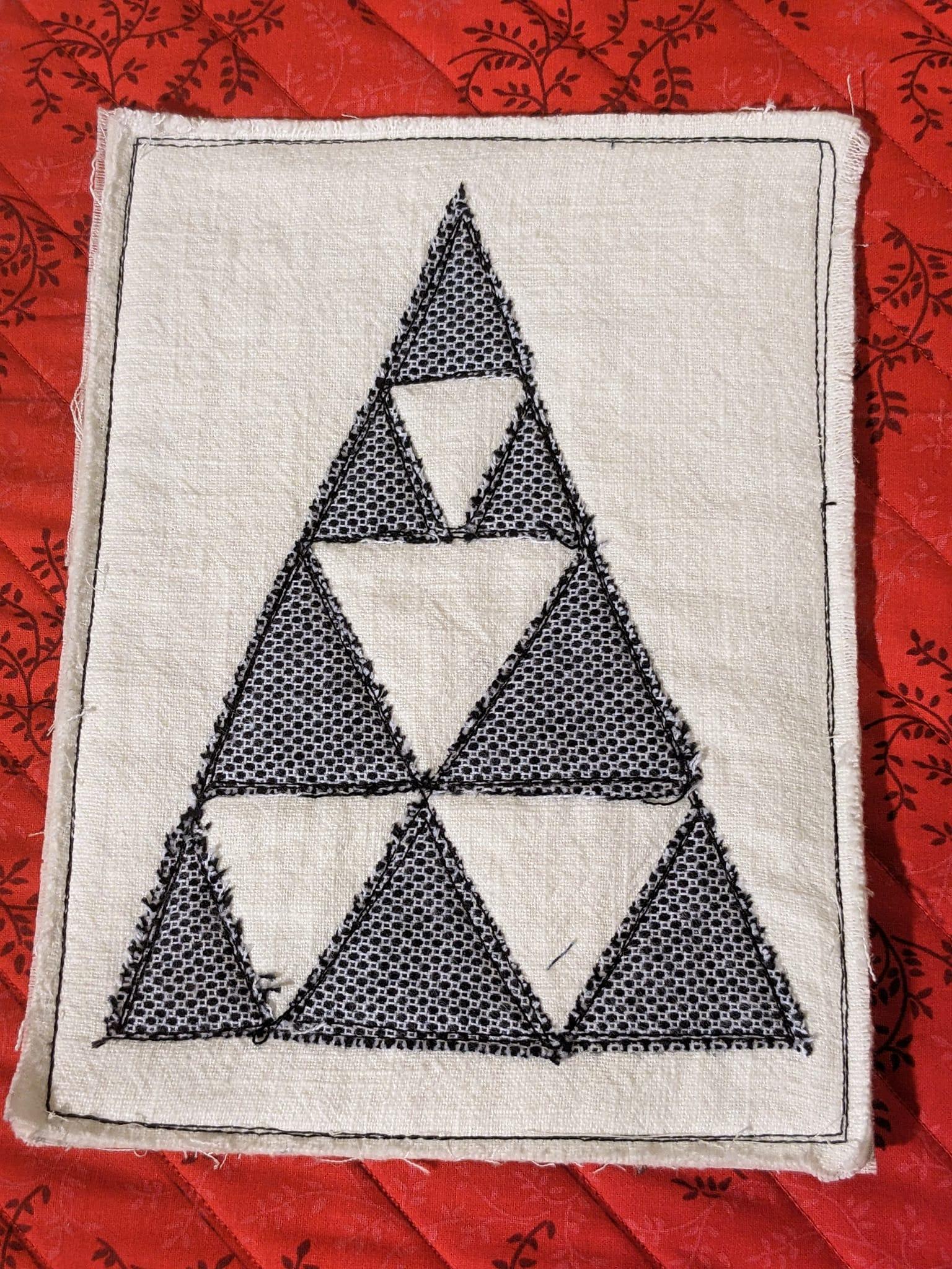 A square quilt with a white background. The quilt features a large, black triangle made up of smaller black triangles. The background is quilted with a subtle, linear pattern.