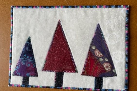 A rectangular quilt with a white background. The quilt features three Christmas trees of different sizes, made of purple and red fabric. The trees have brown trunks. The quilt has a colourful border.