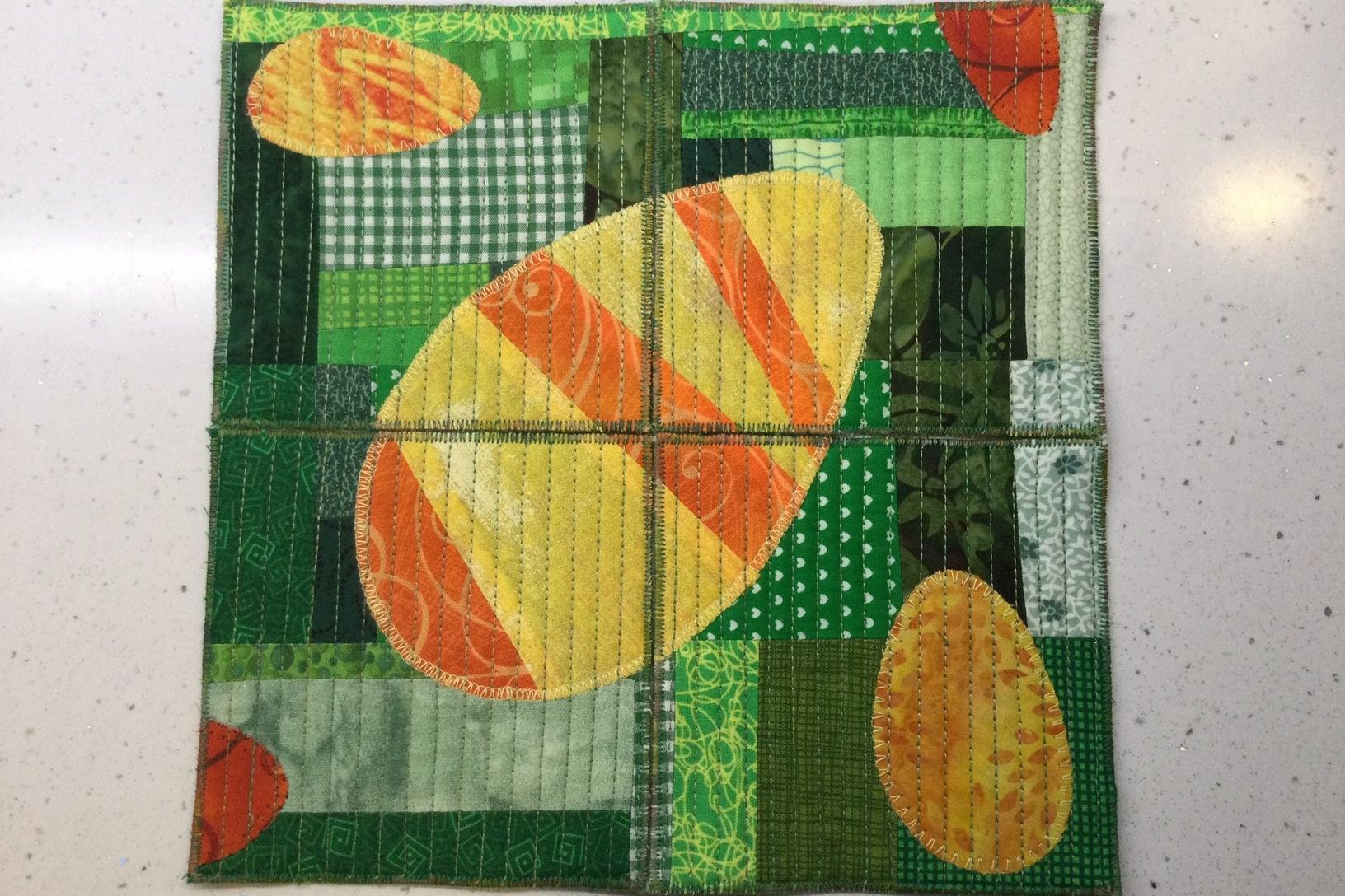 A set of 4 coasters that create a singular image when pieced together. The coasters features three orange and yellow eggs, each with a different pattern and texture. The eggs are surrounded by a patchwork of green fabric in various shades and patterns.