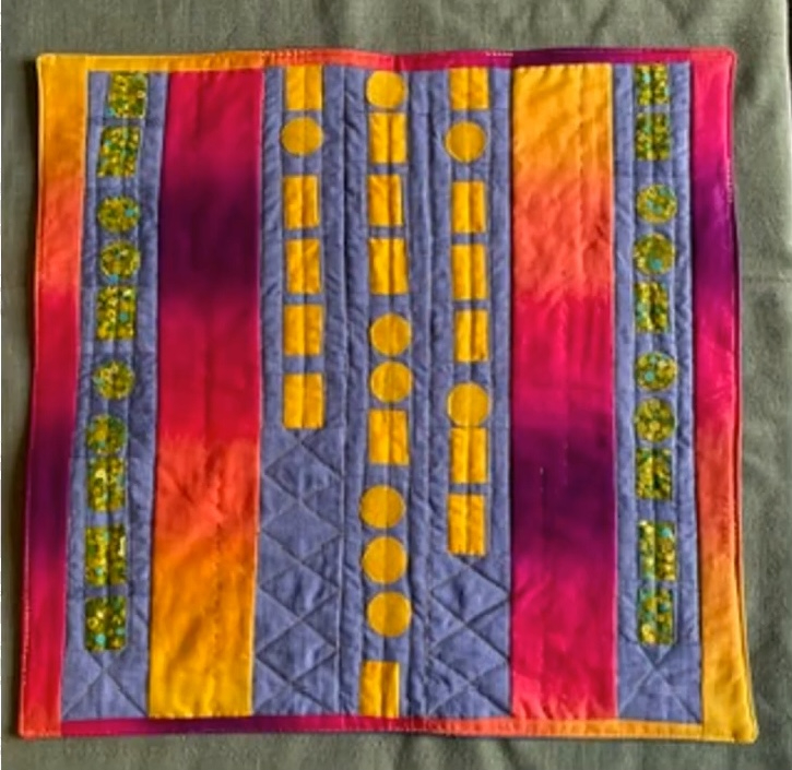A rectangular quilt with a vibrant colour scheme. The quilt features vertical stripes in shades of pink, orange, and yellow. Within these stripes are various shapes, including circles, squares, and rectangles, in a contrasting colour.