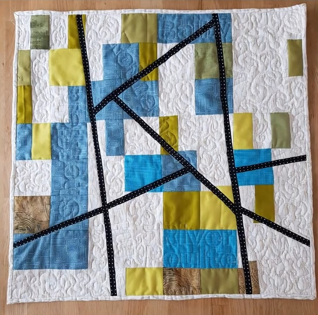 A square quilt with a white background. The quilt features a colorful, geometric design with squares and rectangles in shades of blue, green, and yellow. Black lines intersect the shapes, creating a dynamic composition. The background is quilted with a linear pattern.