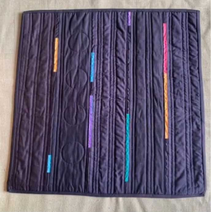 A rectangular quilt with a dark black background. The quilt features vertical, colourful lines in orange, pink, and blue, along with circular shapes. The background is quilted with a linear pattern.