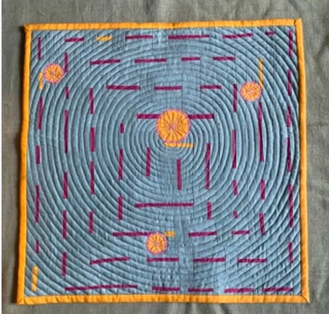 A rectangular quilt with a blue background. The quilt features a circular design with concentric circles in shades of purple. There are also yellow circles scattered throughout. The background is quilted with a linear pattern. The quilt has an orange border.