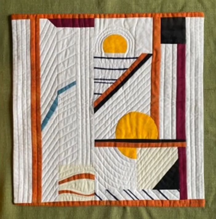 A square quilt with a white background. The quilt features a colorful, geometric design with shapes in orange, yellow, blue, black, and red. The shapes are arranged in a seemingly random pattern, creating a dynamic and visually interesting composition. The background is quilted with a linear pattern. 