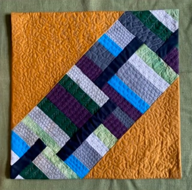 A square quilt with an orange background. The quilt features a diagonal band of colorful rectangles in shades of green, blue, purple, and gray. The background is quilted with a textured, swirling pattern.