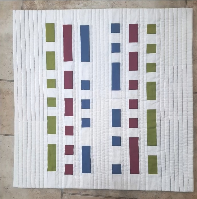 A rectangular quilt with a white background. The quilt features vertical lines of colored rectangles in green, blue, and red. The background is quilted with a subtle linear pattern.