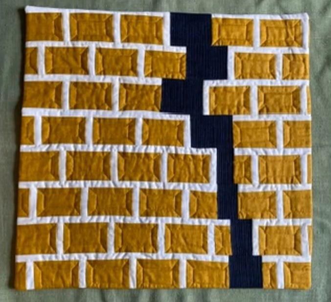 A rectangular quilt with a yellow background. The quilt features a brick pattern, with yellow bricks and white mortar. A black line cuts through the brick pattern, creating a visual divide. The background is quilted with a linear pattern.