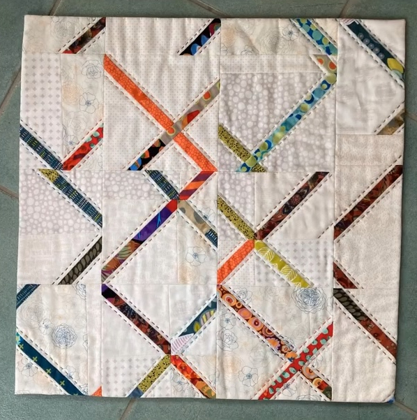 A rectangular quilt with a white background. The quilt features a colorful, geometric design with intersecting lines and shapes in various colors, including orange, blue, and green. The background is quilted with a linear pattern.