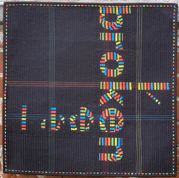 A square quilt with a black background. The word "broken" is spelled out in colorful letters, using strips of fabric in various colours. The background is quilted with a linear pattern. The quilt has a black border.