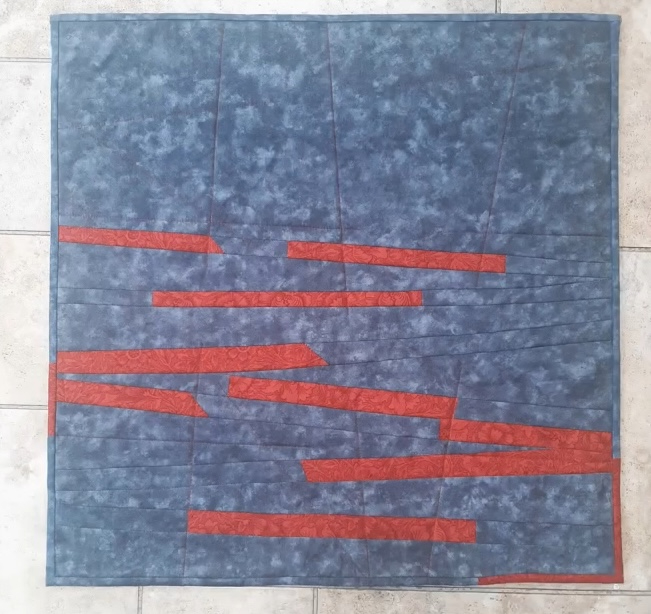 A square quilt with a blue background. The quilt features several horizontal, red strips of varying lengths, arranged in a staggered pattern. The background has a subtle, textured pattern.