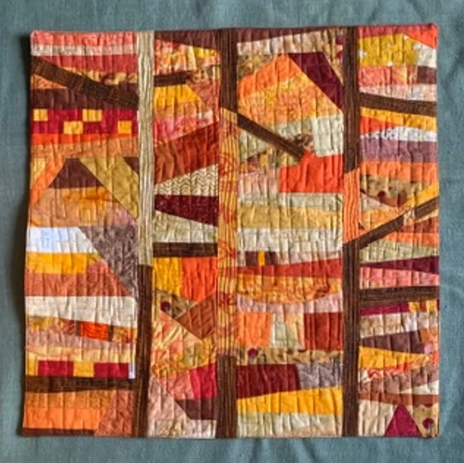 A rectangular quilt with a vibrant, warm color palette. The quilt features a patchwork design with rectangles, triangles, and squares in shades of orange, red, brown, and yellow. The shapes overlap and intersect, creating a dynamic and visually interesting composition. The background is quilted with a linear pattern.