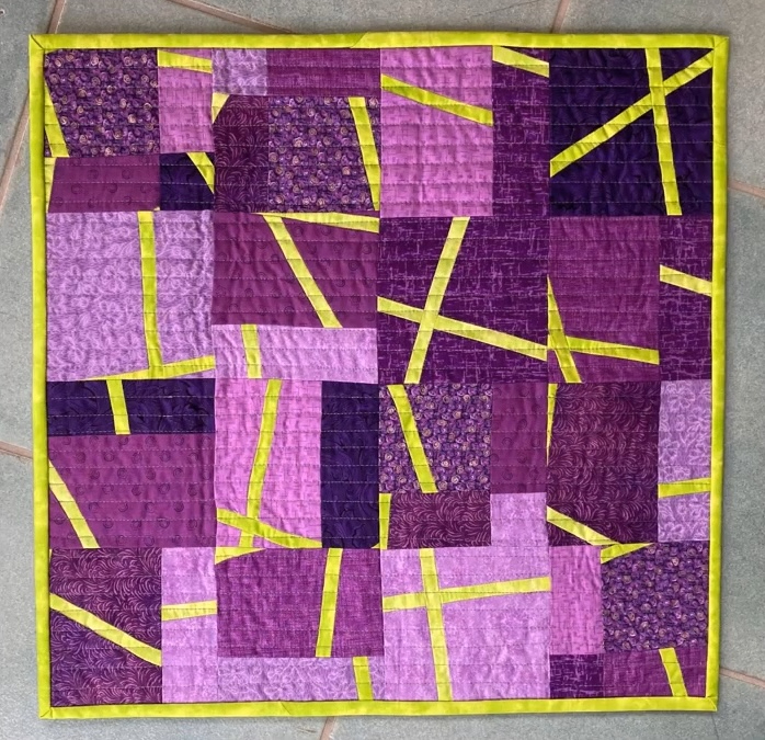 A square quilt with a predominantly purple background. The quilt features a patchwork design with squares and rectangles in various shades of purple. Yellow lines and shapes are overlaid on the purple fabric, creating a geometric pattern. The quilt has a yellow border.