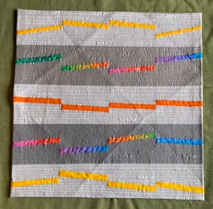 A rectangular quilt with a gray background. The quilt features horizontal bands of color, including yellow, orange, red, pink, purple, and blue. These bands are interrupted by smaller, colorful shapes, creating a dynamic and playful composition. The background is quilted with a linear pattern.