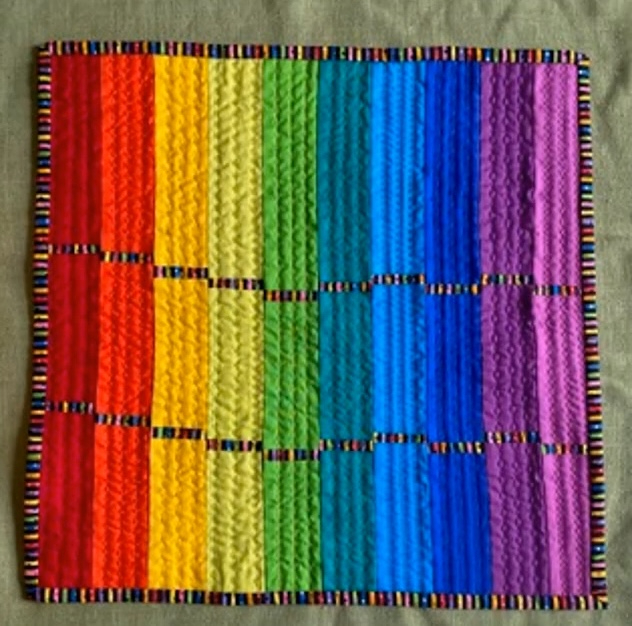 A rectangular quilt with a rainbow-colored design. The quilt features vertical stripes in shades of red, orange, yellow, green, blue, indigo, and violet. The background is quilted with a linear pattern. The quilt has a colorful border.
