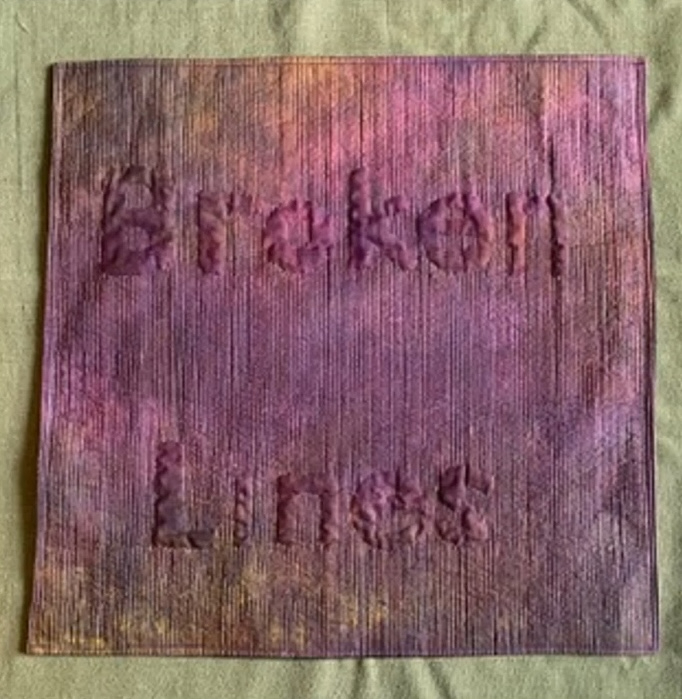 A rectangular quilt with a purple background. The words "Broken Lines" are embroidered in white thread on the fabric. The background has a textured, marbled effect. The quilt has a purple border.