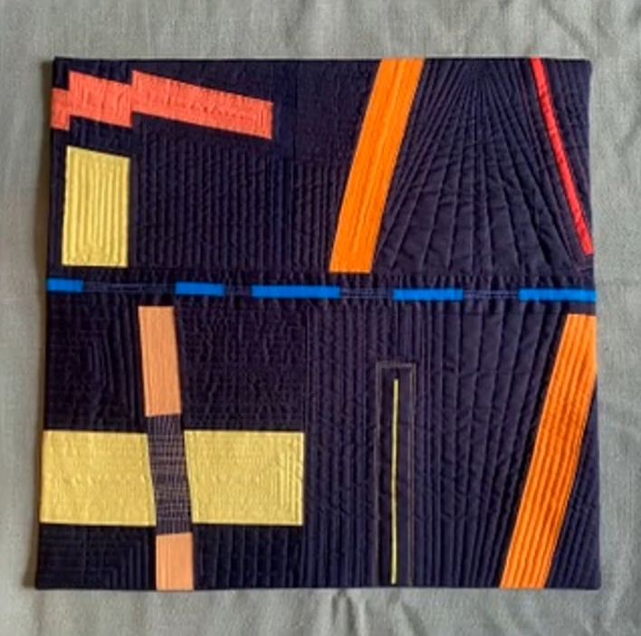 A rectangular quilt with a dark blue background. The quilt features a vibrant, abstract design with geometric shapes in orange, yellow, and red. The shapes are arranged in a seemingly random pattern, creating a dynamic and visually interesting composition. The background is quilted with a linear pattern.