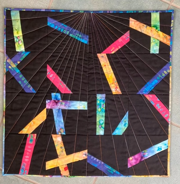A square quilt with a dark background. The quilt features colorful, rectangular strips radiating outwards from a central point. The strips are in various colors, including pink, orange, yellow, green, and blue. The background is quilted with radiating lines. 