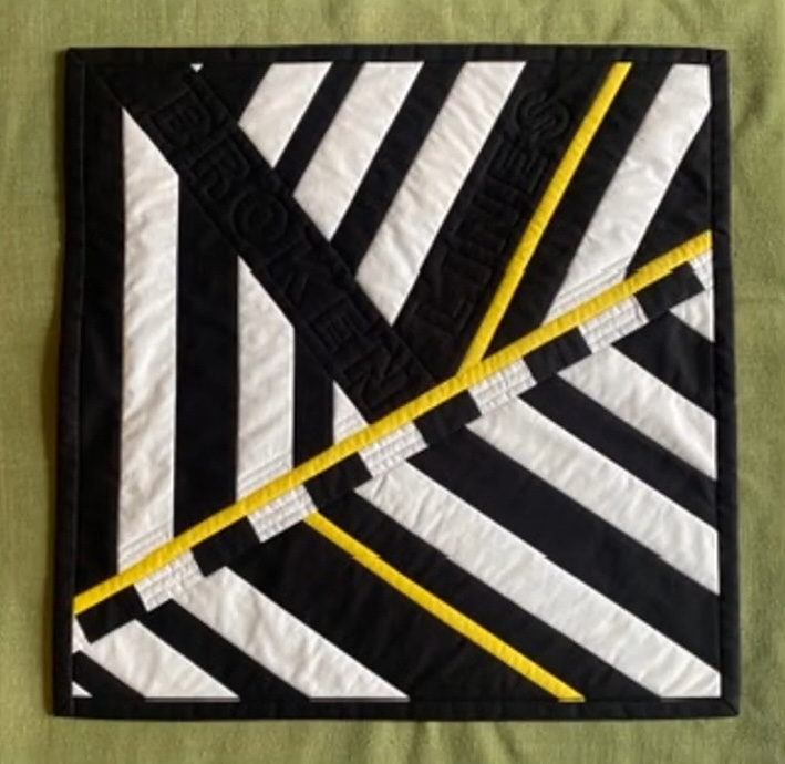 A square quilt with a black background. The quilt features a geometric design with diagonal stripes in black and white. The word "Broken Lines" is spelled out in yellow letters across the black fabric. The quilt has a black border.