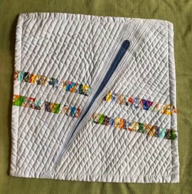 A rectangular quilt with a white background. The quilt features a blue, vertical line cutting through the center, with a colorful, patchwork strip on either side. The background is quilted with a diamond pattern.